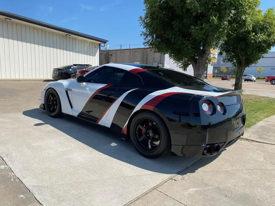 A white, black, and red custom-wrapped Nissan GT-R R35 with aftermarket wheels parked outside a building.