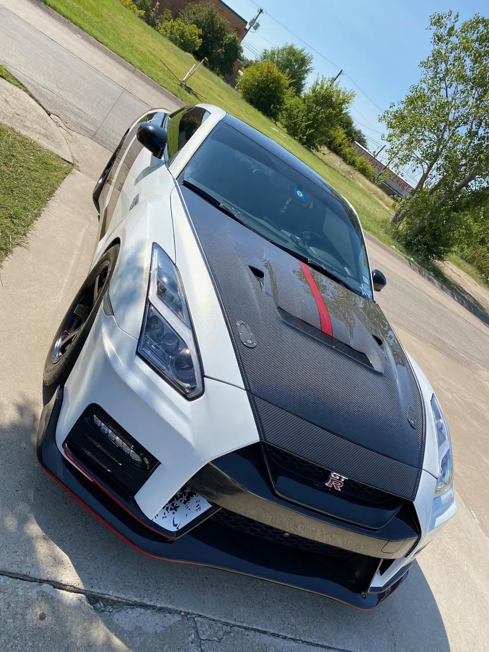 A white Nissan GT-R with a carbon fiber hood and red stripe parked on a sunny driveway.