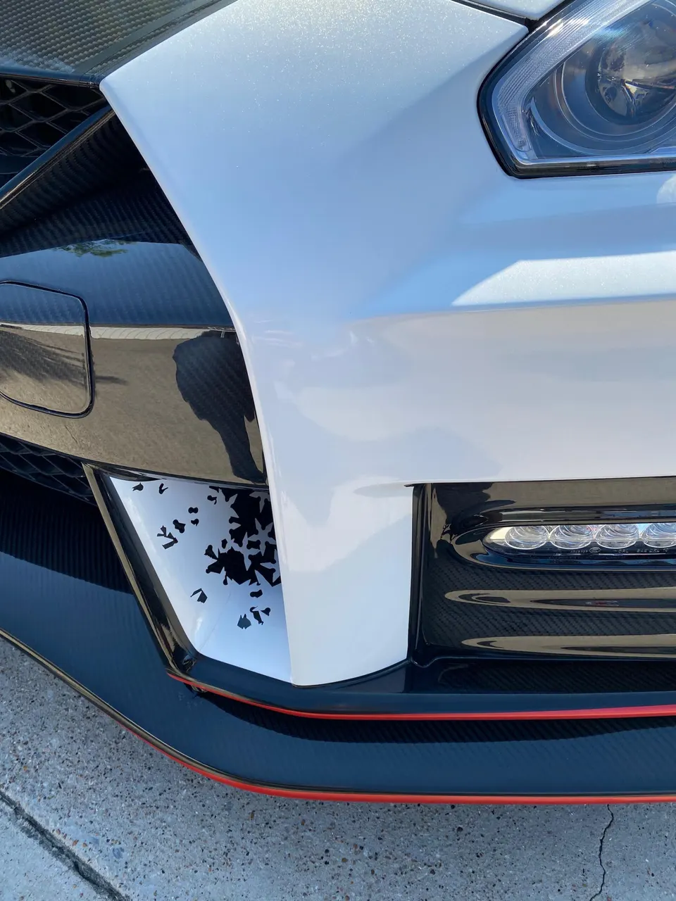 Close-up of a white Nissan GT-R front bumper with carbon fiber splitter, custom decal, and LED daytime running lights.