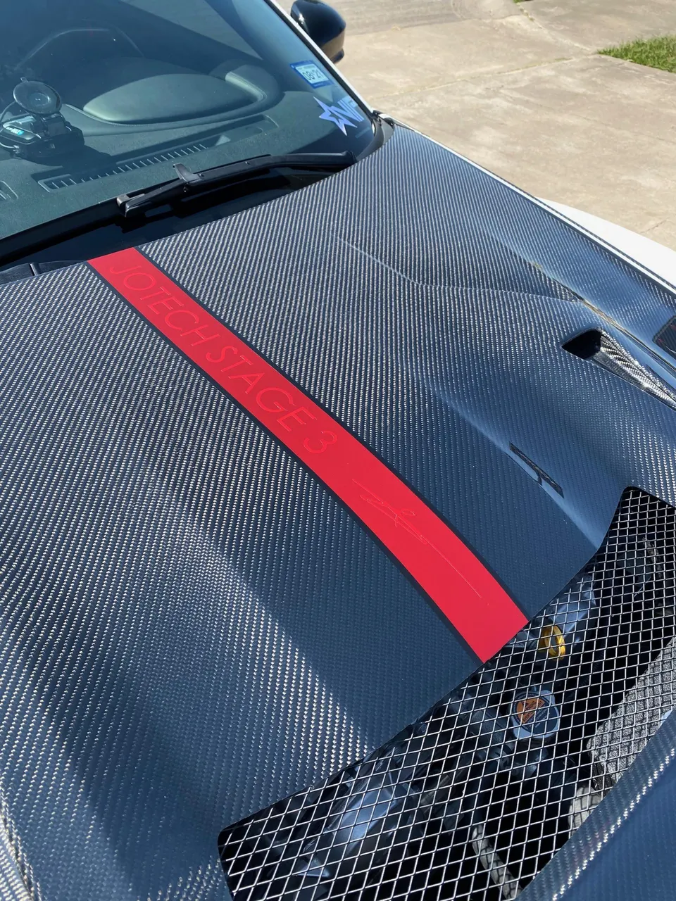 Close-up of a car's carbon fiber hood with a red Jotech Stage 3 decal and a mesh hood vent.