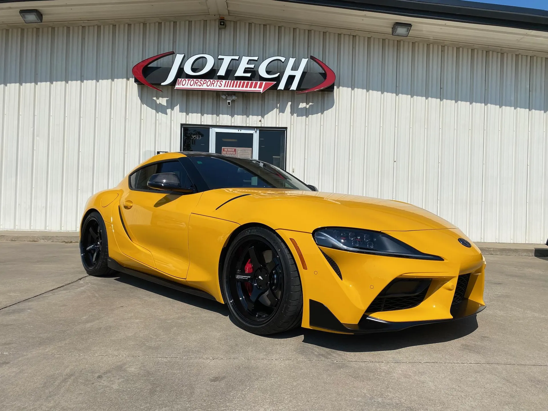 A yellow Toyota Supra with aftermarket wheels parked in front of the Jotech Motorsports building.
