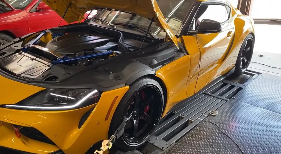 Yellow Toyota Supra on a dynamometer with its hood open, undergoing performance testing at a tuning shop.