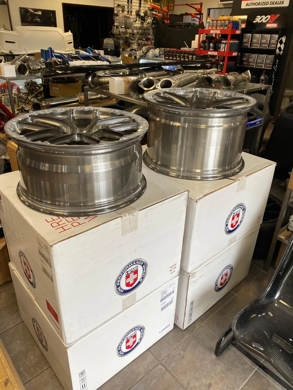 New HRE performance wheels on boxes in a tuning shop, with other car parts and shelves in the background.