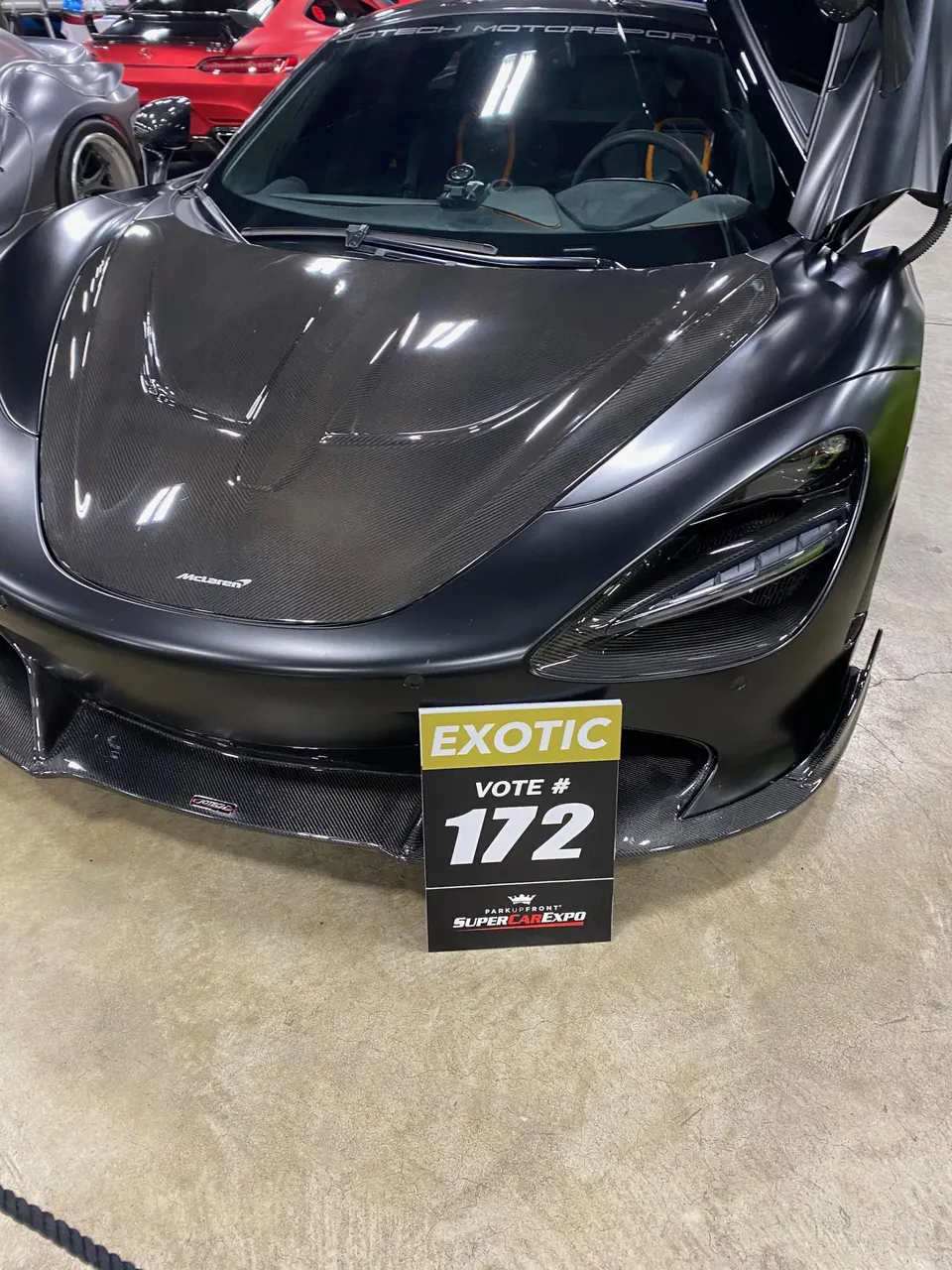 Matte black McLaren supercar with carbon fiber hood and front splitter displayed at a car show.