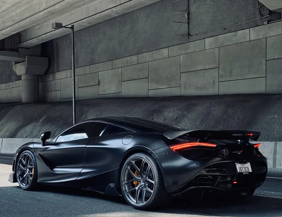 Matte black McLaren 720S with aftermarket wheels and a large rear wing parked under a concrete overpass.