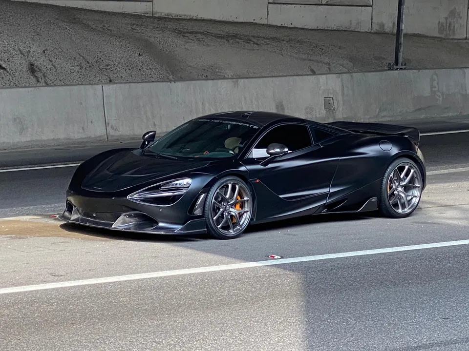 A black McLaren 720S with aftermarket wheels and carbon fiber aero parked on a road under an overpass.
