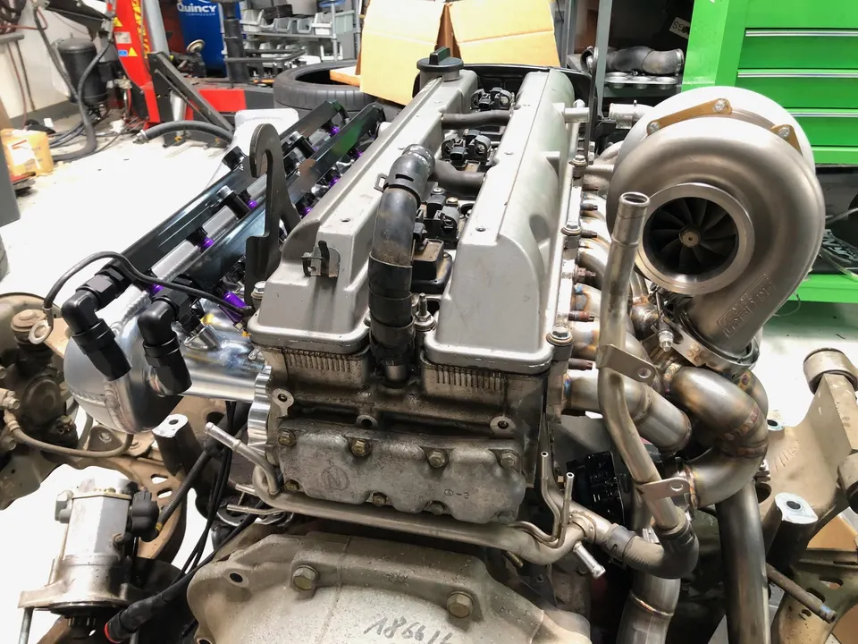 Close-up of a high-performance engine with a large turbocharger, aftermarket intake manifold, and fuel rails in a shop.