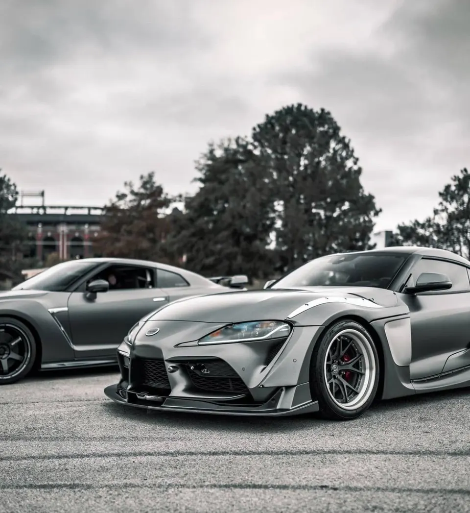 Modified grey Toyota Supra and Nissan GT-R parked outdoors, showcasing custom widebody kits and aftermarket wheels.