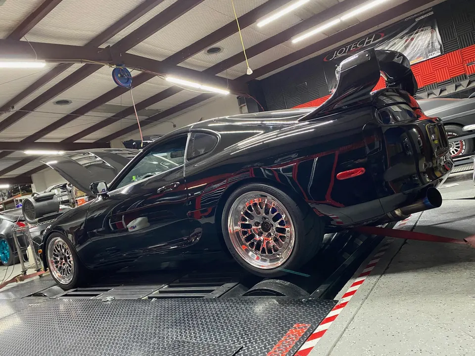 A black Toyota Supra MK4 on a dyno at a performance tuning shop, showcasing aftermarket wheels and a large rear wing.