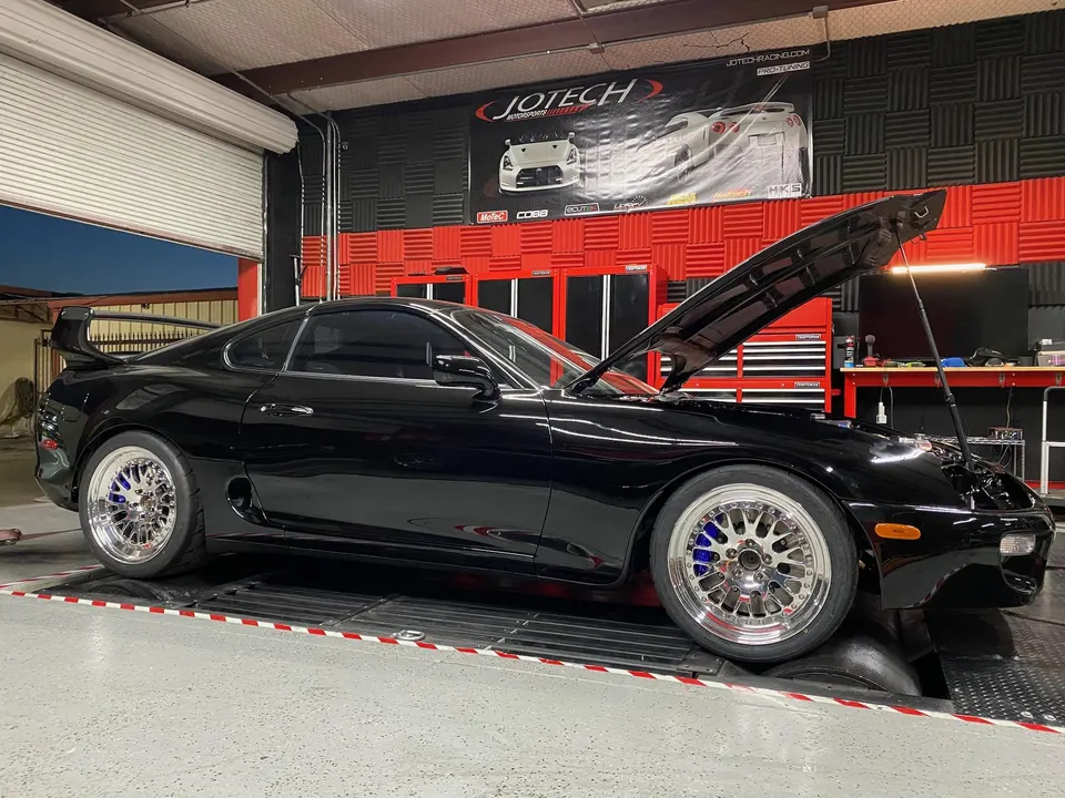 Black Toyota Supra MK4 on a dynamometer at Jotech Motorsports with the hood open, showcasing aftermarket wheels.