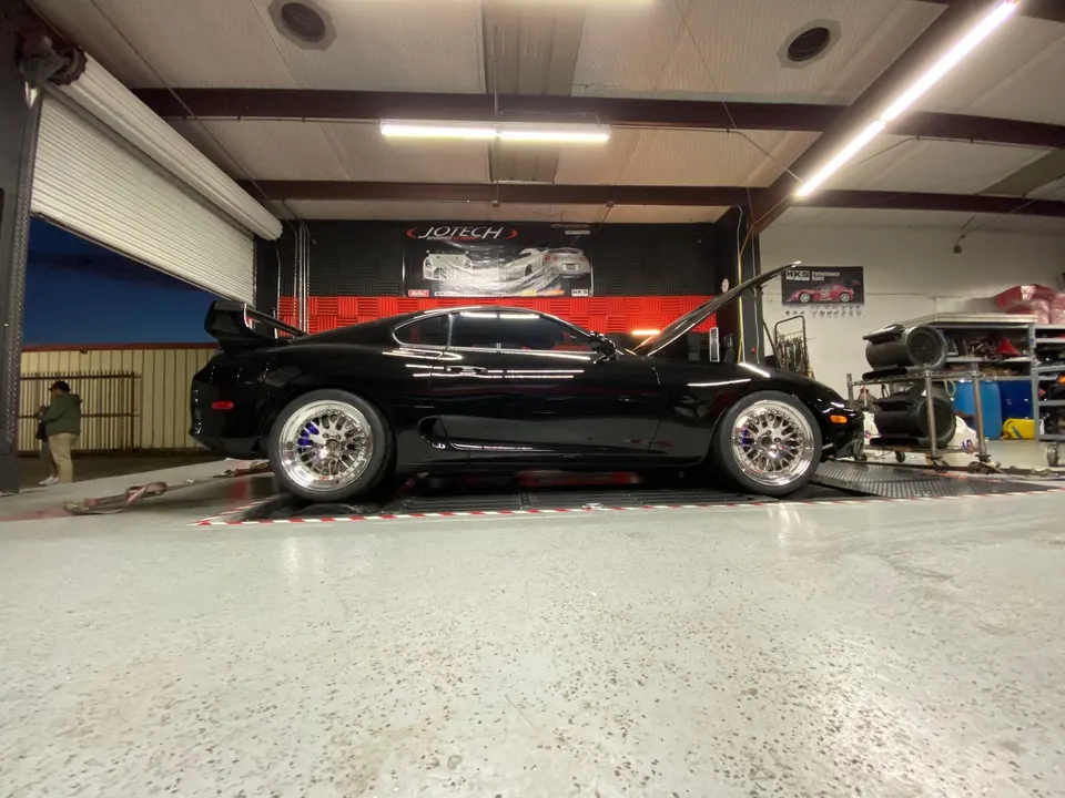 A black Toyota Supra on a dyno at Jotech Motorsports, undergoing performance tuning in a shop setting.