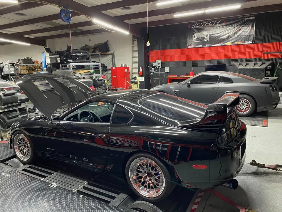Black Toyota Supra MKIV on a dyno with its hood open, next to a grey Nissan GT-R in a performance tuning shop.