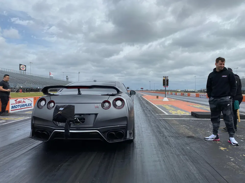 A gray Nissan GT-R with a parachute attached to the rear, waiting at the staging lane of a drag strip under a cloudy sky.