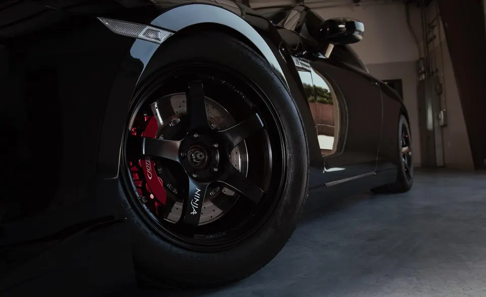 A close-up of a black Nissan GT-R's front wheel with black multi-spoke rims and red Jotech brake calipers.