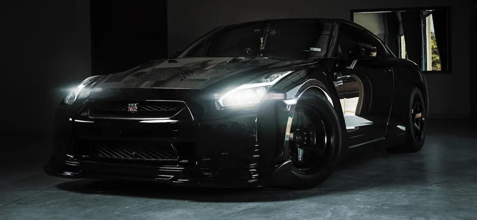 A sleek black Nissan GT-R with a carbon fiber hood and bright headlights, parked in a dark studio setting.