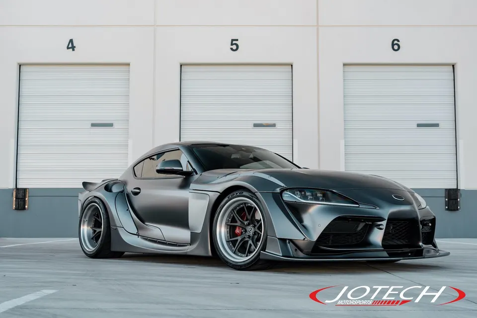 A matte grey widebody Toyota Supra with aftermarket wheels parked in front of garage doors.