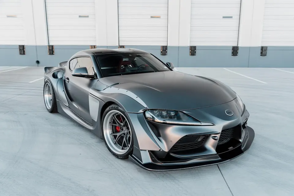 A custom matte grey widebody Toyota Supra with aftermarket wheels and aero modifications parked in a modern setting.