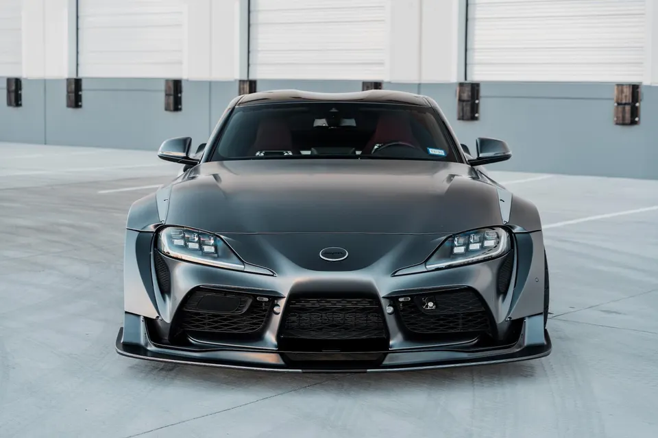 A front view of a grey widebody Toyota Supra A90 with an aggressive front splitter parked in a clean industrial setting.