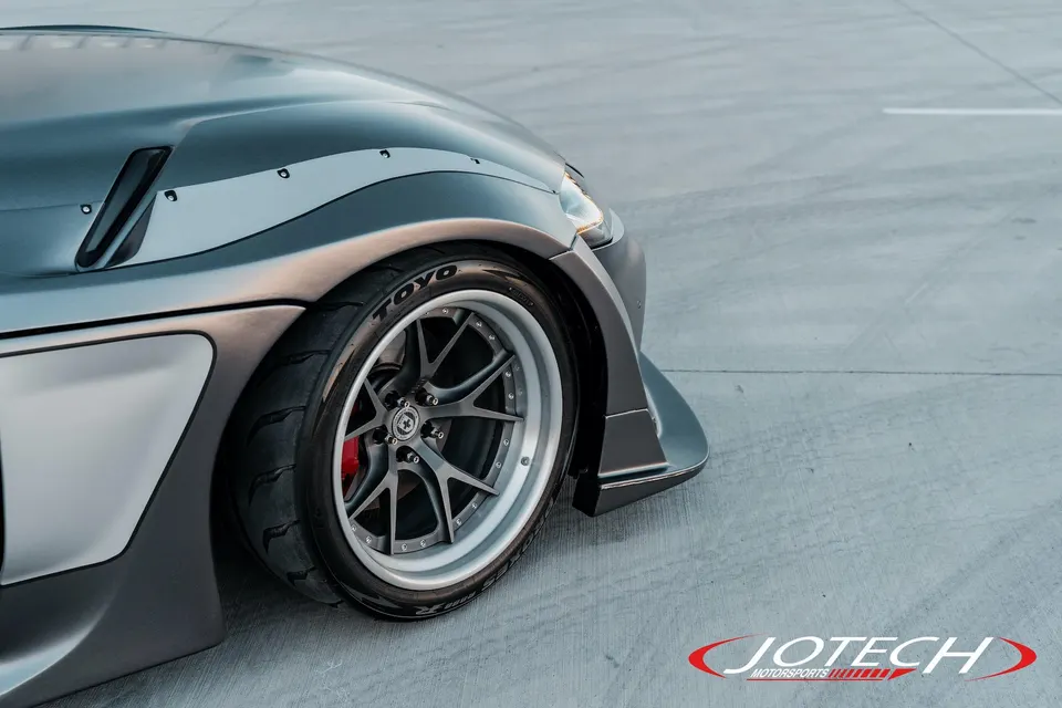Close-up of a grey widebody car's front wheel, featuring custom multi-spoke wheels, Toyo tires, and red brake calipers.