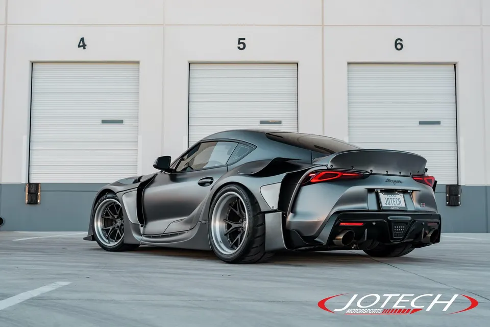 Matte grey widebody Toyota Supra with aftermarket wheels and a large rear wing parked in front of garage doors.