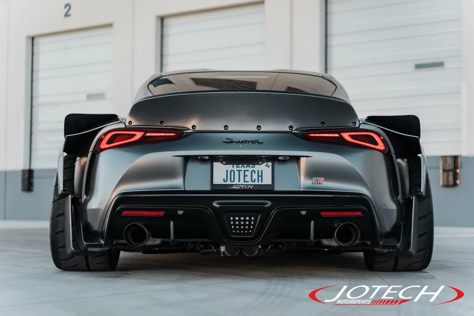 Rear view of a grey widebody Toyota Supra with a large rear wing and aftermarket exhaust, parked in front of garage doors.