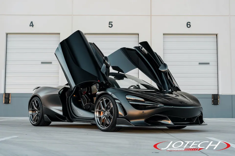 Black McLaren 720S with butterfly doors open, parked in front of white garage doors.