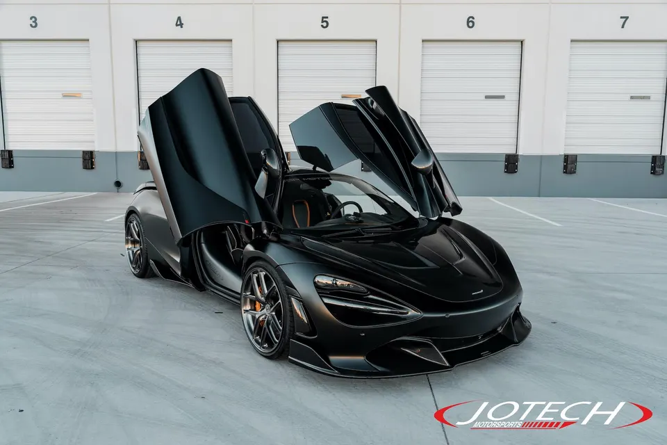 A black McLaren 720S with its butterfly doors open, parked in front of white garage doors, showcasing its sleek design.