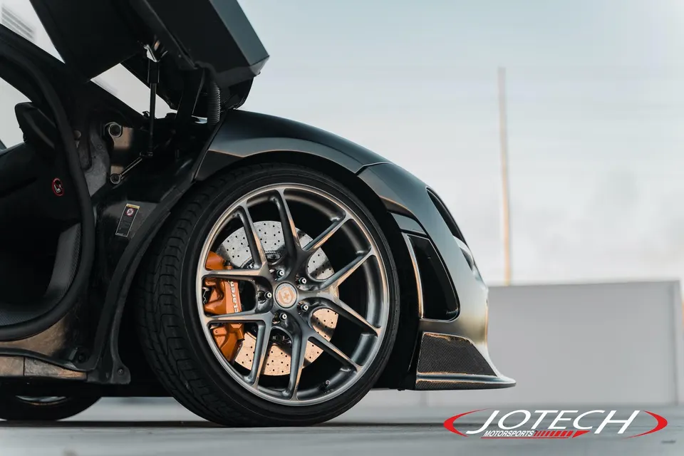 A close-up of a black McLaren's front wheel with HRE wheels and orange McLaren brake calipers, featuring carbon fiber aero.