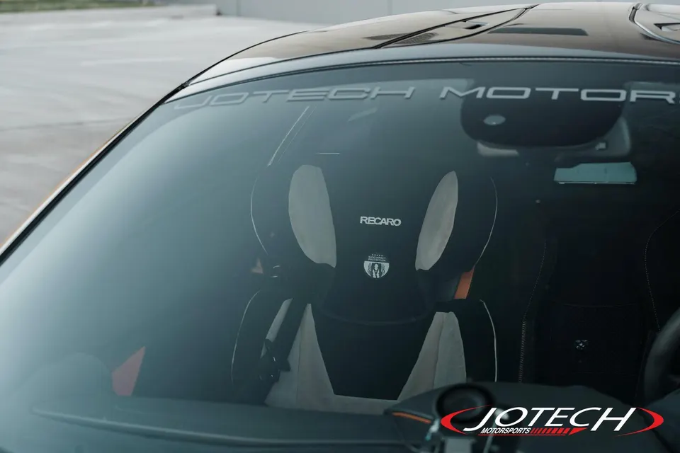 A Recaro child seat visible through the windshield of a Jotech Motorsports car, with the Jotech logo on the glass.