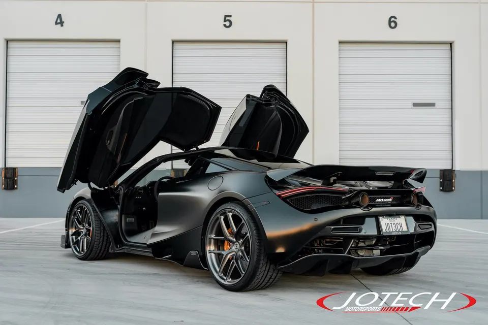 Matte black McLaren 720S with butterfly doors open, parked in front of garage doors. Custom wheels and rear diffuser visible.