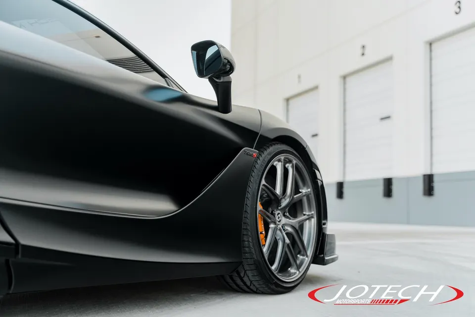 Close-up of a black McLaren 720S showcasing custom wheels with orange brake calipers and a sleek side mirror.