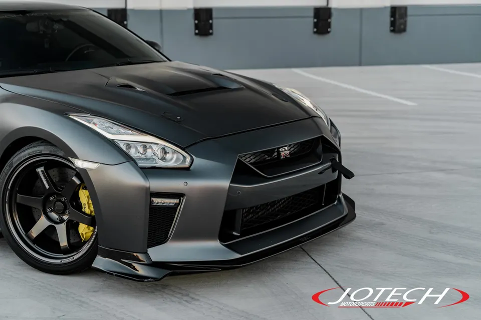 Matte grey Nissan GT-R with custom hood, front splitter, aftermarket wheels, and yellow brake calipers parked in a lot.