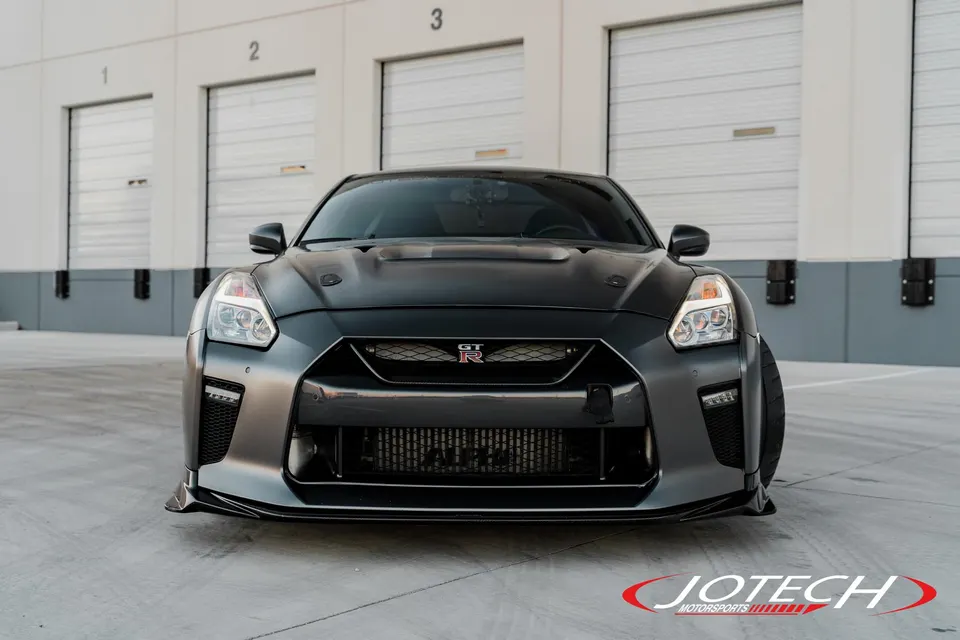 Front view of a dark grey Nissan GT-R R35 with an aftermarket front splitter and large intercooler parked in front of a warehouse.