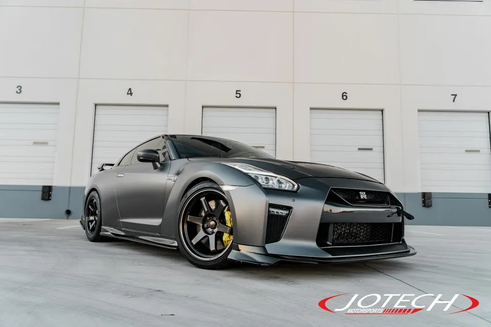 Matte grey Nissan GT-R R35 with aftermarket wheels and a front splitter parked in front of garage doors.