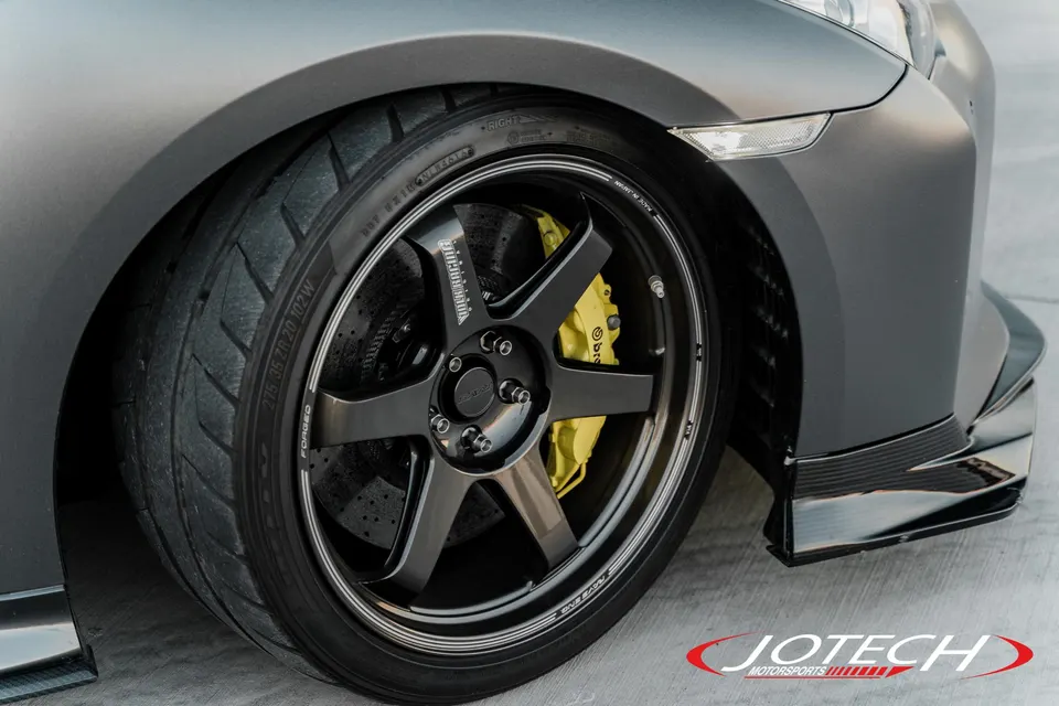 A close-up of a Nissan GT-R's front wheel, featuring black forged wheels, yellow Brembo calipers, and performance tires.