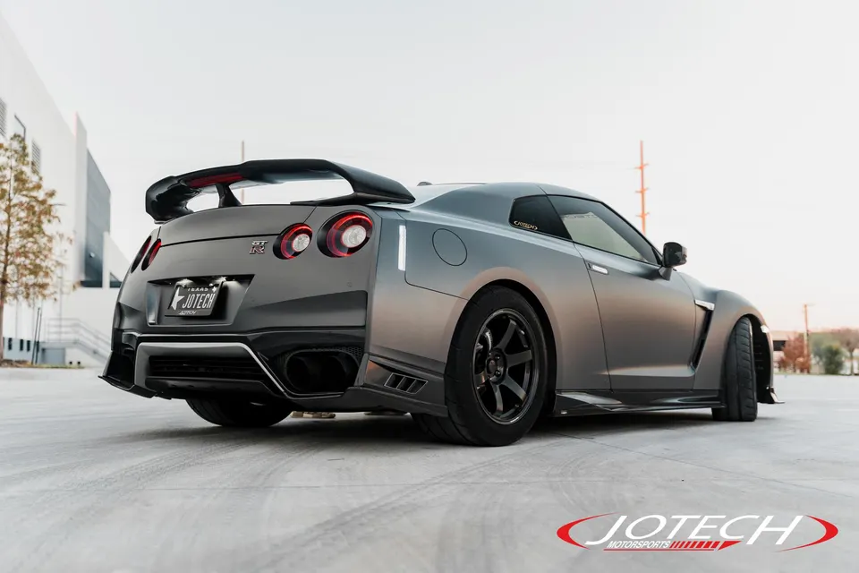 A custom gray Nissan GT-R with a widebody kit, large rear wing, and aftermarket wheels parked outdoors.