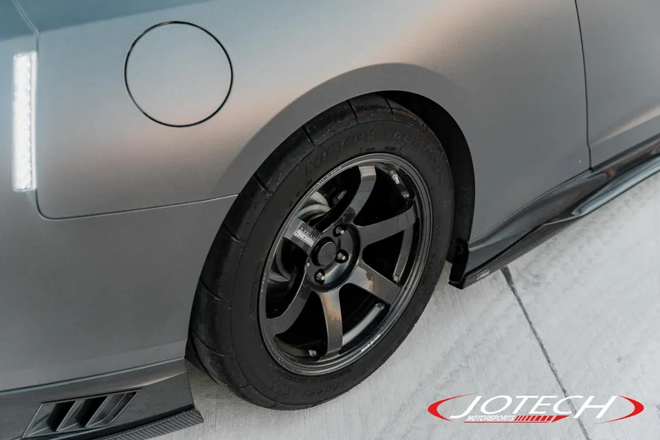 A close-up of a matte gray Nissan GT-R's rear wheel, featuring black Rays TE37 wheels and a carbon fiber side skirt.