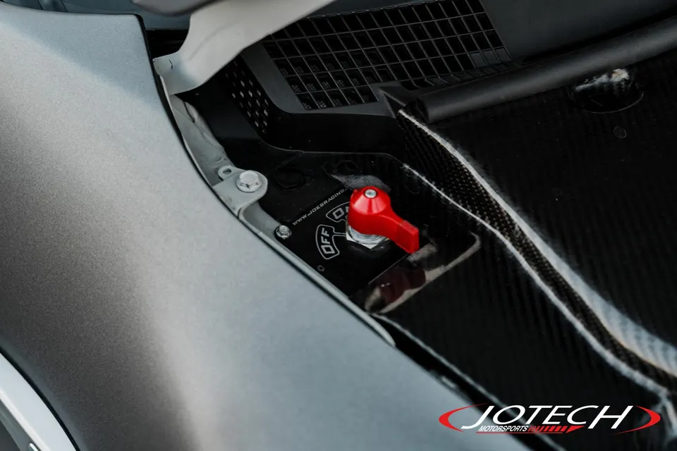 A close-up of a red battery disconnect switch installed in a car's engine bay with carbon fiber trim.