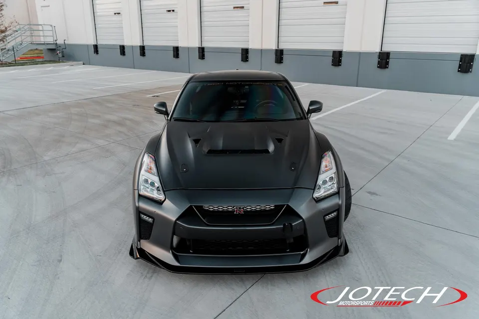 Front view of a matte grey Nissan GT-R R35 with an aftermarket hood and front splitter, parked in an industrial lot.