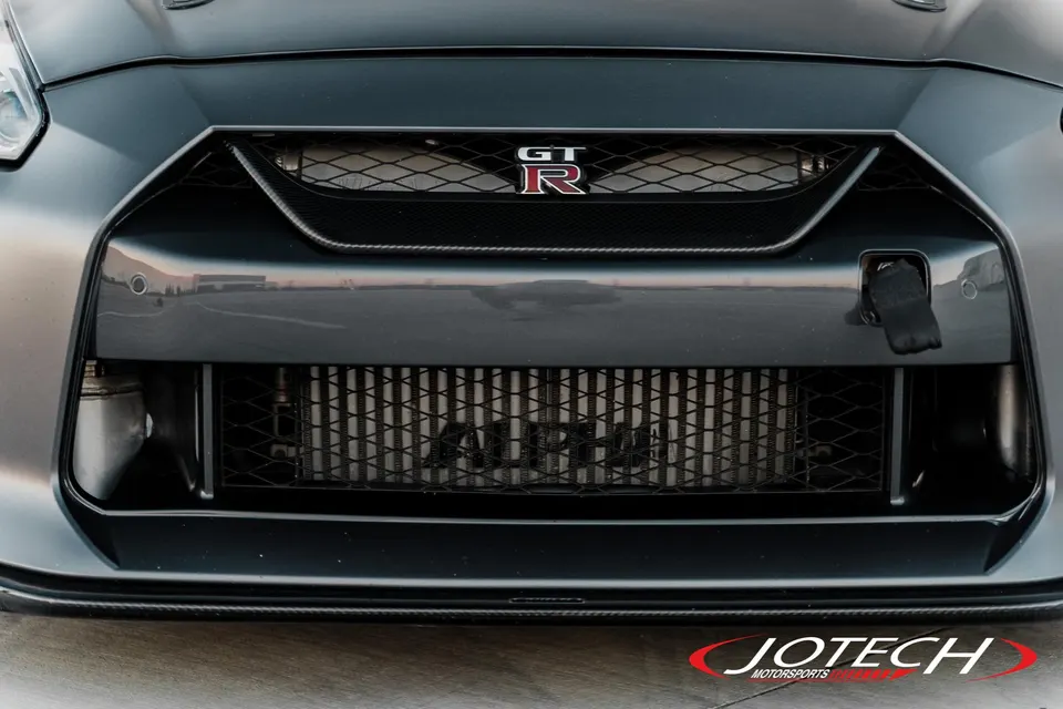 Close-up of the front bumper of a grey Nissan GT-R, showcasing an aftermarket Alpha Performance intercooler and GT-R badge.