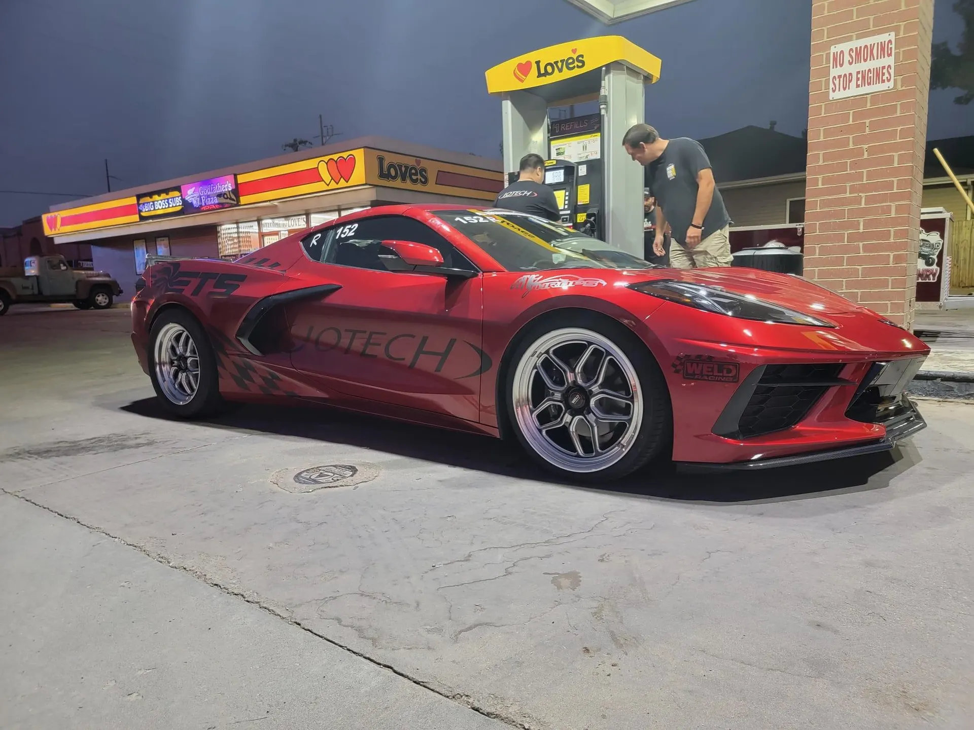 A red C8 Corvette with Jotech livery refueling at a Love's gas station at night, with two men standing by the pump.