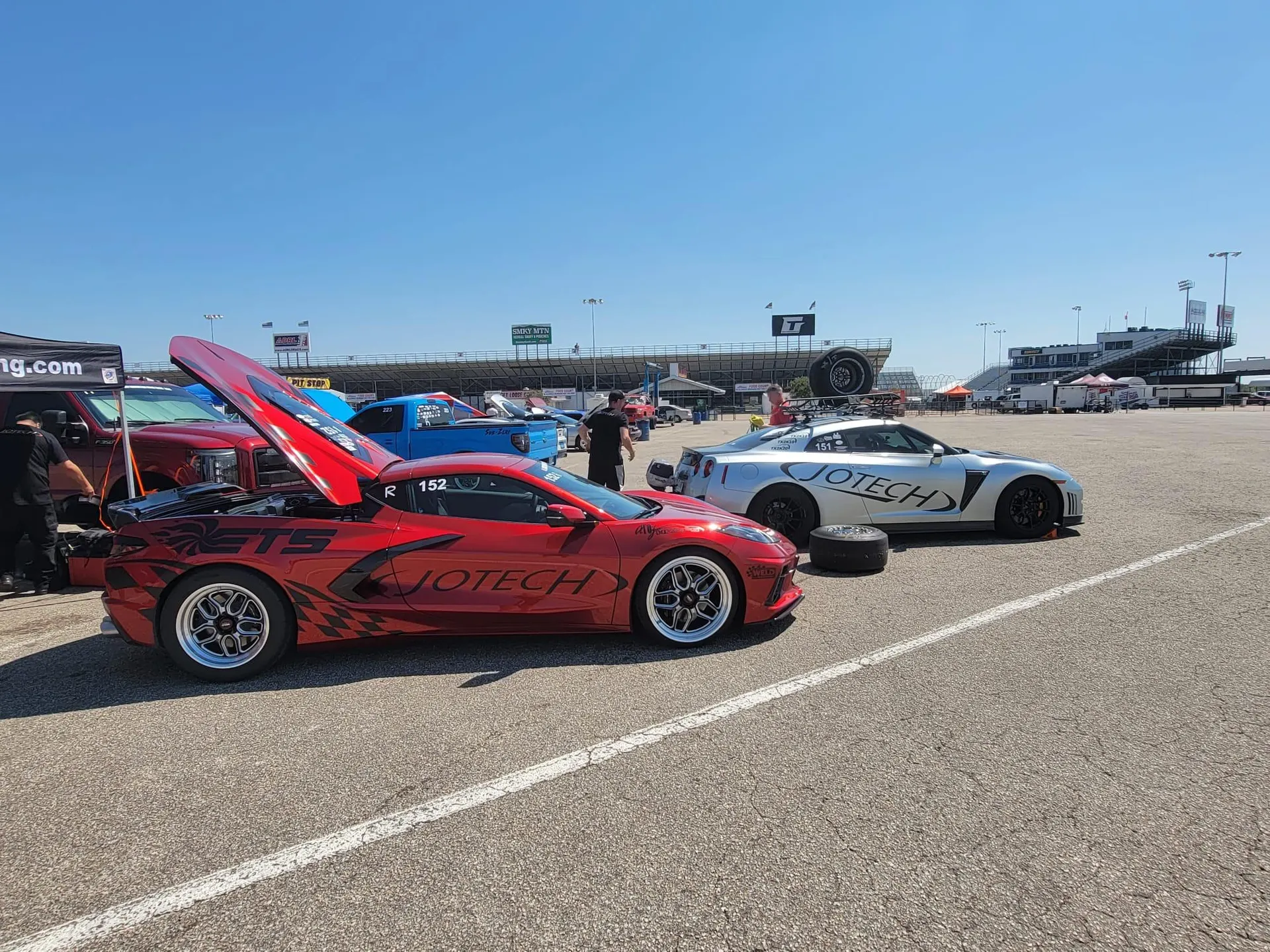 Red Corvette C8 and silver Nissan GT-R race cars with Jotech branding parked at a drag strip paddock on a sunny day.