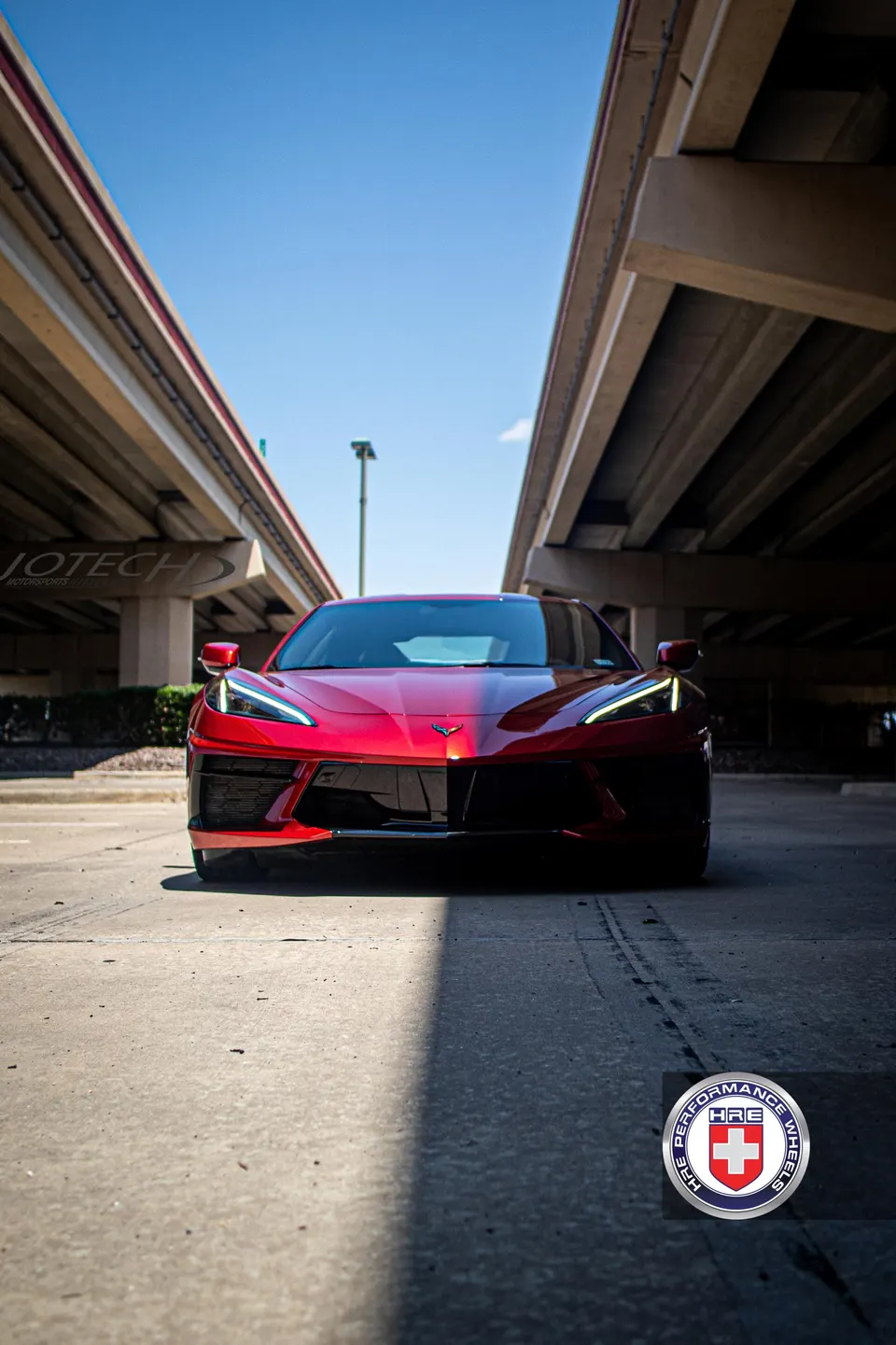 A low-angle front view of a red Chevrolet Corvette C8 parked under an overpass on a sunny day.