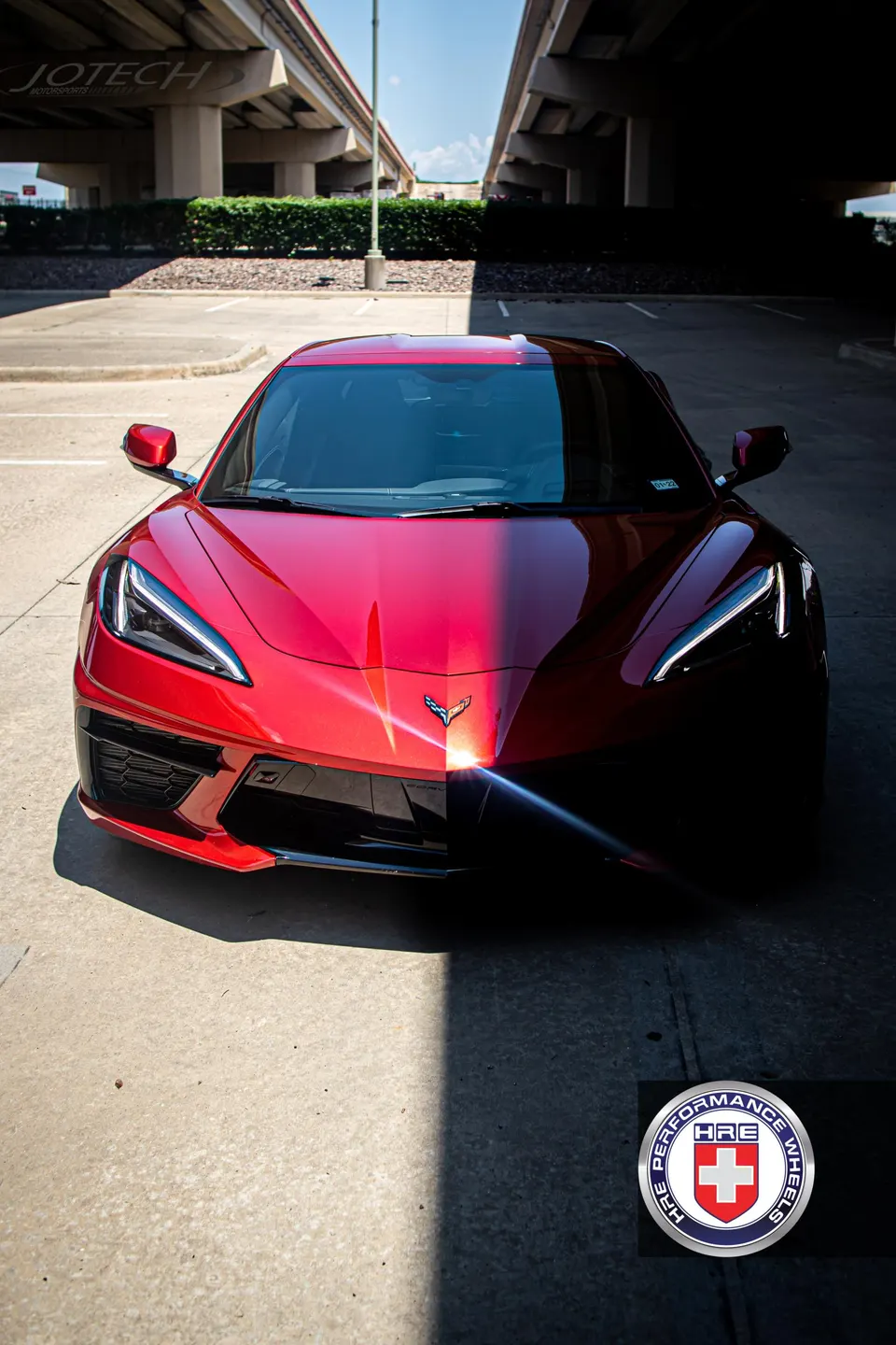 A striking red Corvette C8 parked outdoors, with its front half dramatically split between sunlight and shadow, under highway overpasses.