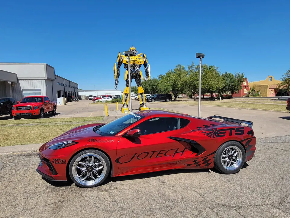 A red Corvette C8.R with custom drag wheels and Jotech Motorsports livery parked outdoors next to a large yellow robot statue.
