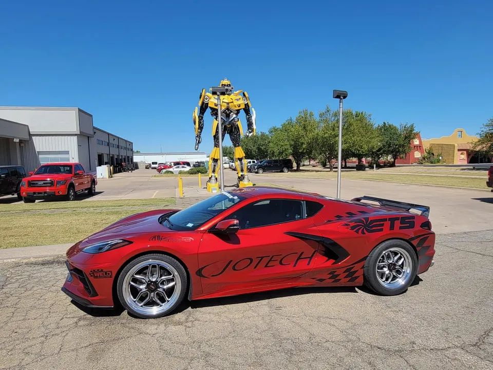 Red C8 Corvette with Jotech graphics and drag wheels parked outdoors next to a large yellow robot statue.