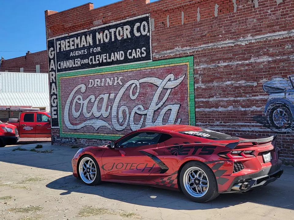 Red Corvette C8.R with drag wheels and custom livery parked next to an old brick building with a Coca-Cola mural.