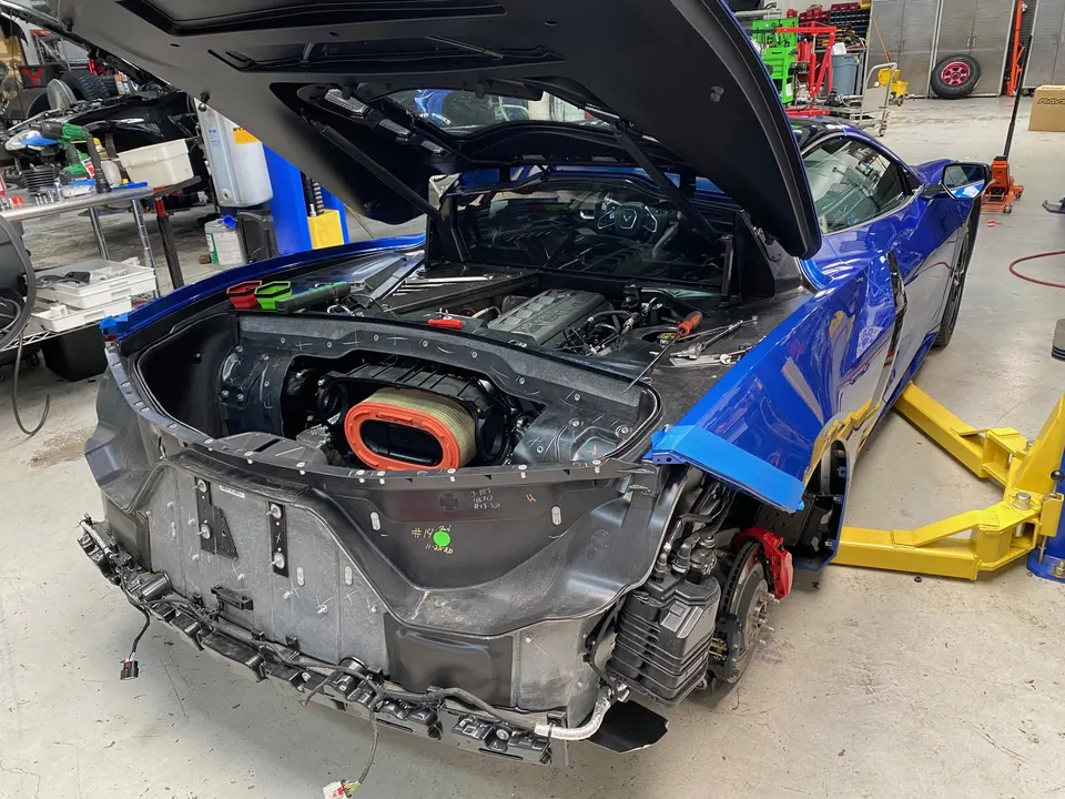 A blue Corvette C8 with its rear bumper removed, exposing the engine bay, air filter, and rear suspension on a shop lift.