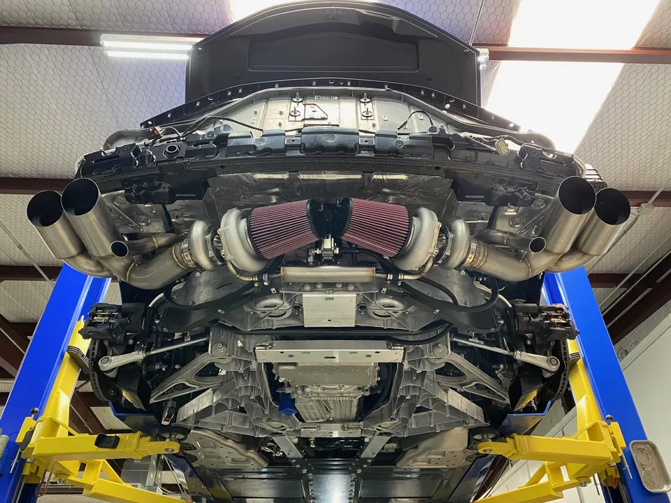 Underside view of a C8 Corvette on a lift, showcasing a custom twin-turbo system with large air filters and quad exhaust tips.