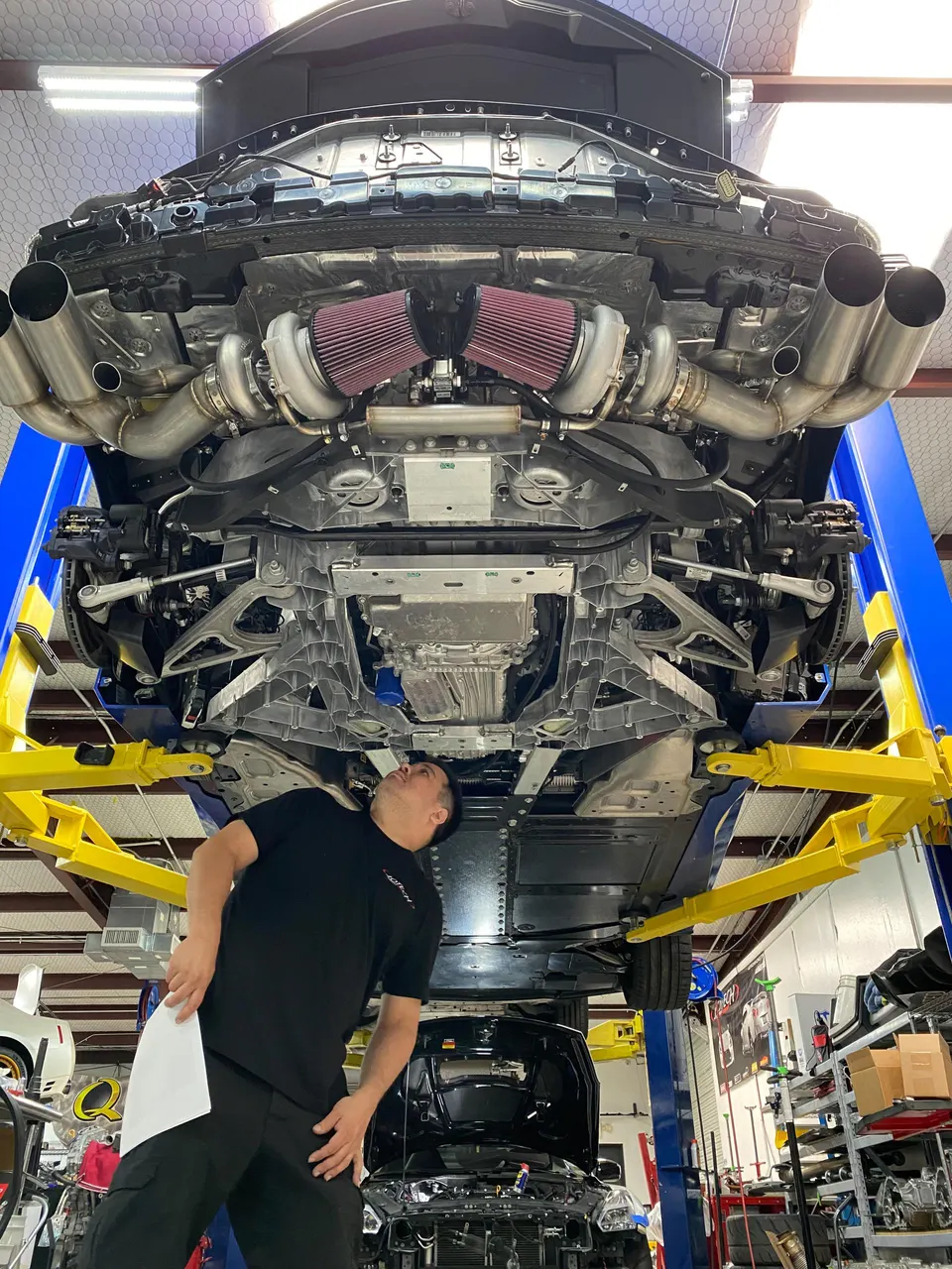Underside of a performance car on a lift, showing twin turbos, red air filters, and custom exhaust. Mechanic inspects.